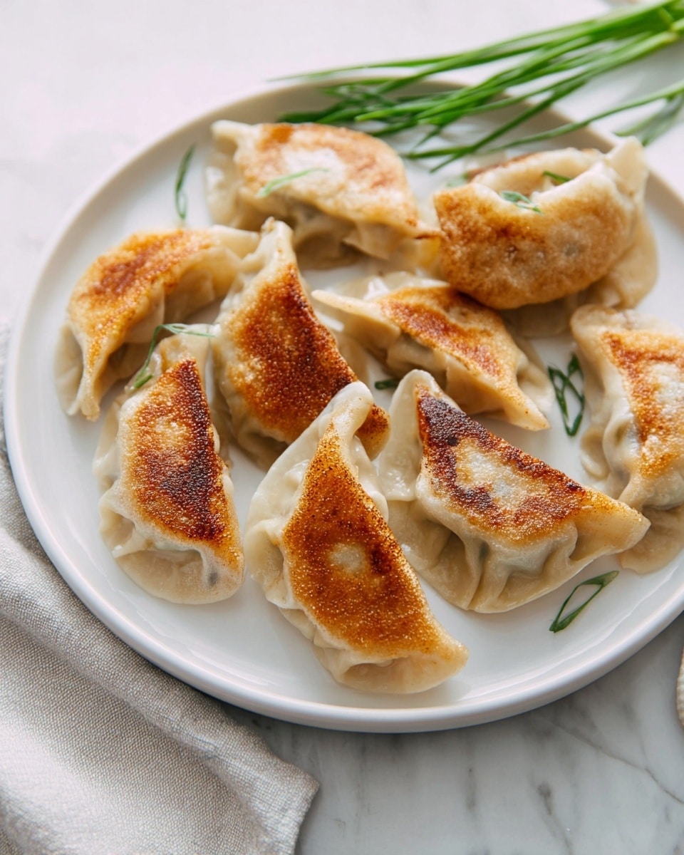 An oval white plate holds eleven pan-fried dumplings arranged in a loose circular pattern, each dumpling showing a golden brown, crispy bottom side and a pale, steamed dough top side with delicate folds. To the top left of the plate, a small white bowl with intricate colorful patterns is filled with dark reddish soy dipping sauce speckled with chili flakes. Next to the bowl, there is a decorative pink teapot with white and blue details. Fresh green chives lay beside a white textured cloth in the bottom left corner. Light wooden chopsticks rest on the white marbled surface to the bottom right of the plate. The photo is taken with an iphone --ar 4:5 --v 7