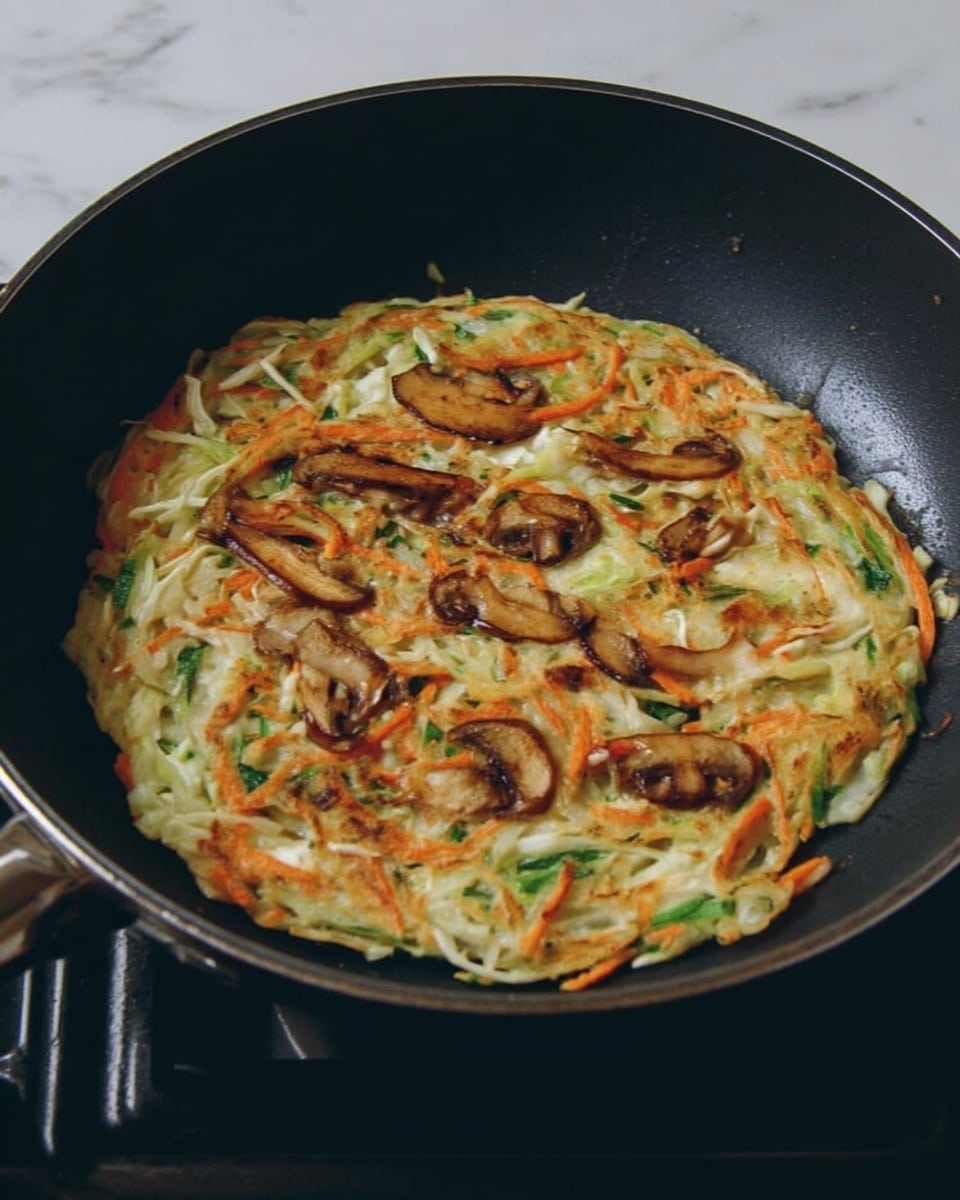 In a black pan on a stove, there is a round pancake made with mixed vegetables. The pancake has a light golden color with visible thin strips of orange carrots, green beans, white cabbage, and browned mushroom slices on top. The texture looks crispy on the edges and soft in the middle, with the vegetables spread evenly. The background shows the stovetop with black grates. photo taken with an iphone --ar 4:5 --v 7