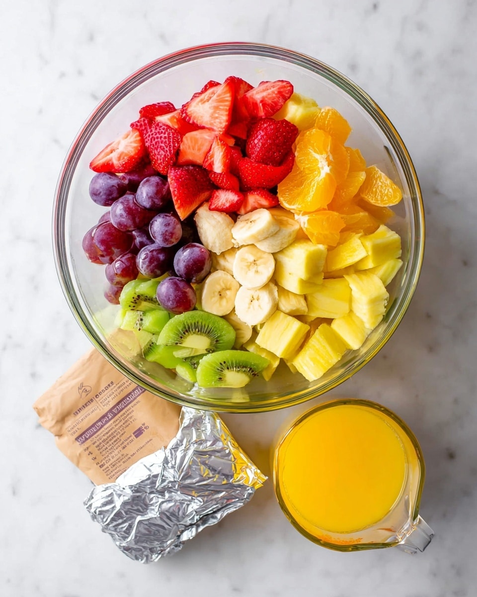 A clear glass bowl sits on a white marbled surface filled with six sections of assorted fresh fruit, arranged side by side in a circular pattern. Starting from the top left going clockwise, the sections include bright red sliced strawberries, yellow cubed pineapple, small orange segments, green sliced kiwi, red whole and halved grapes, and yellow banana slices layered over the center of the bowl. Below the bowl on the surface, there is a block of silver wrapped cheese, a brown paper bag, and a glass measuring cup filled with orange juice. The photo is taken with an iphone --ar 4:5 --v 7